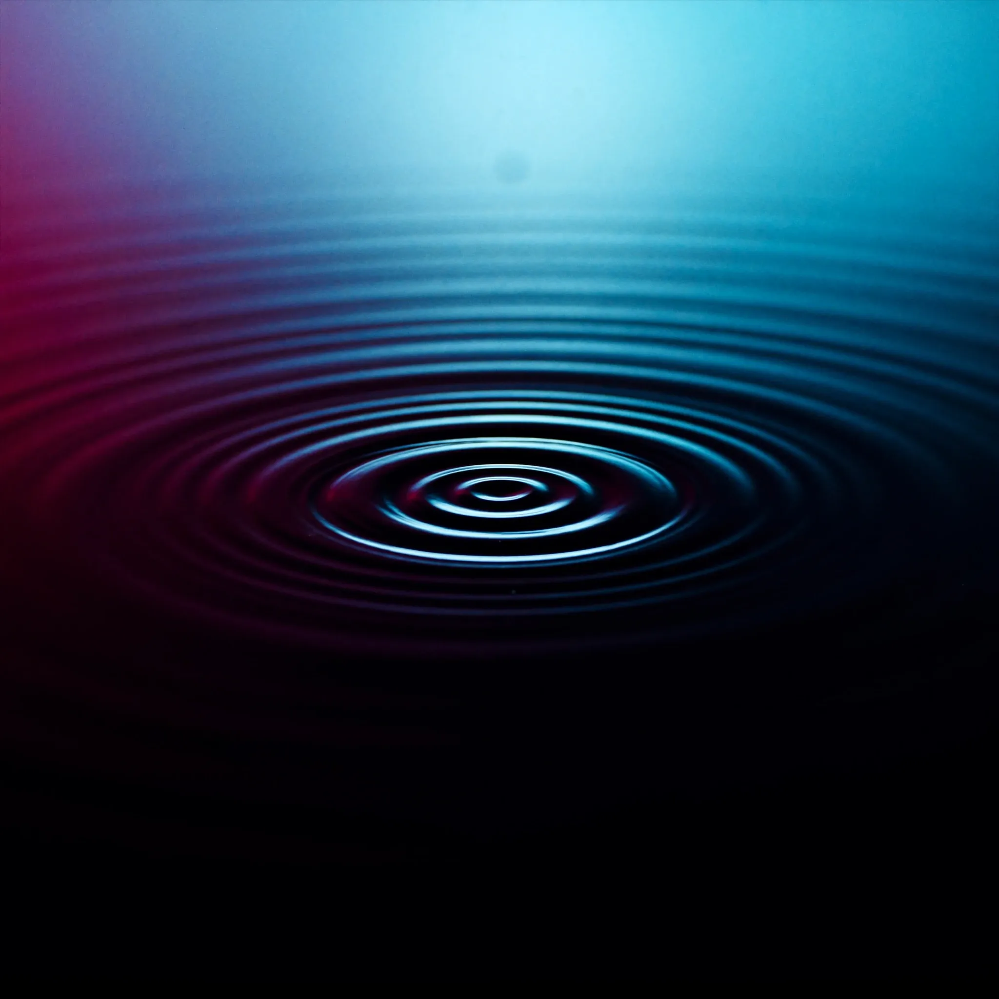 A single ripple spreading in perfect concentric rings across a still black pool, lit cyan from above
