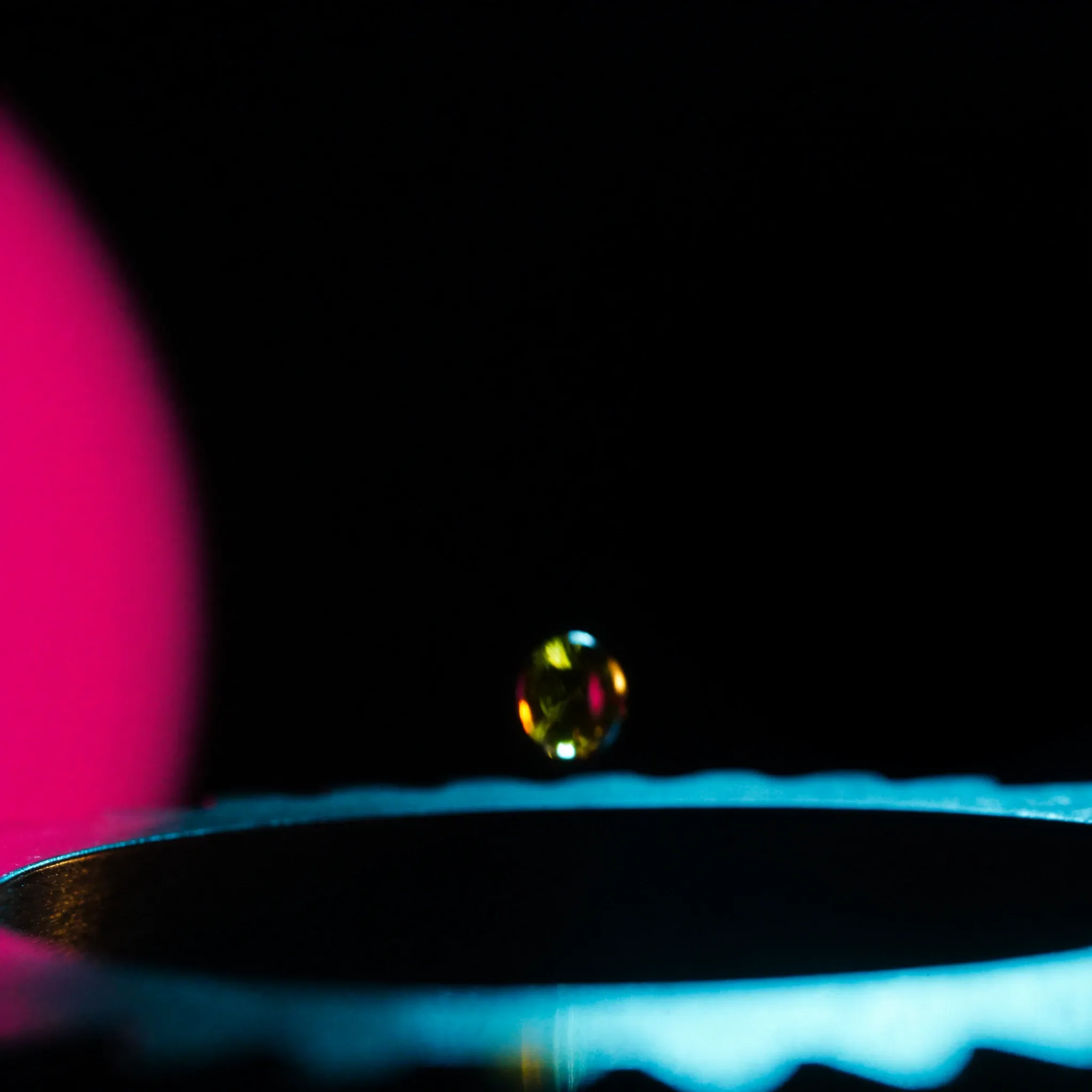 A single drop of golden oil falling onto the teeth of a polished steel gear, macro, cyan rim light, the gear clearly visible against black