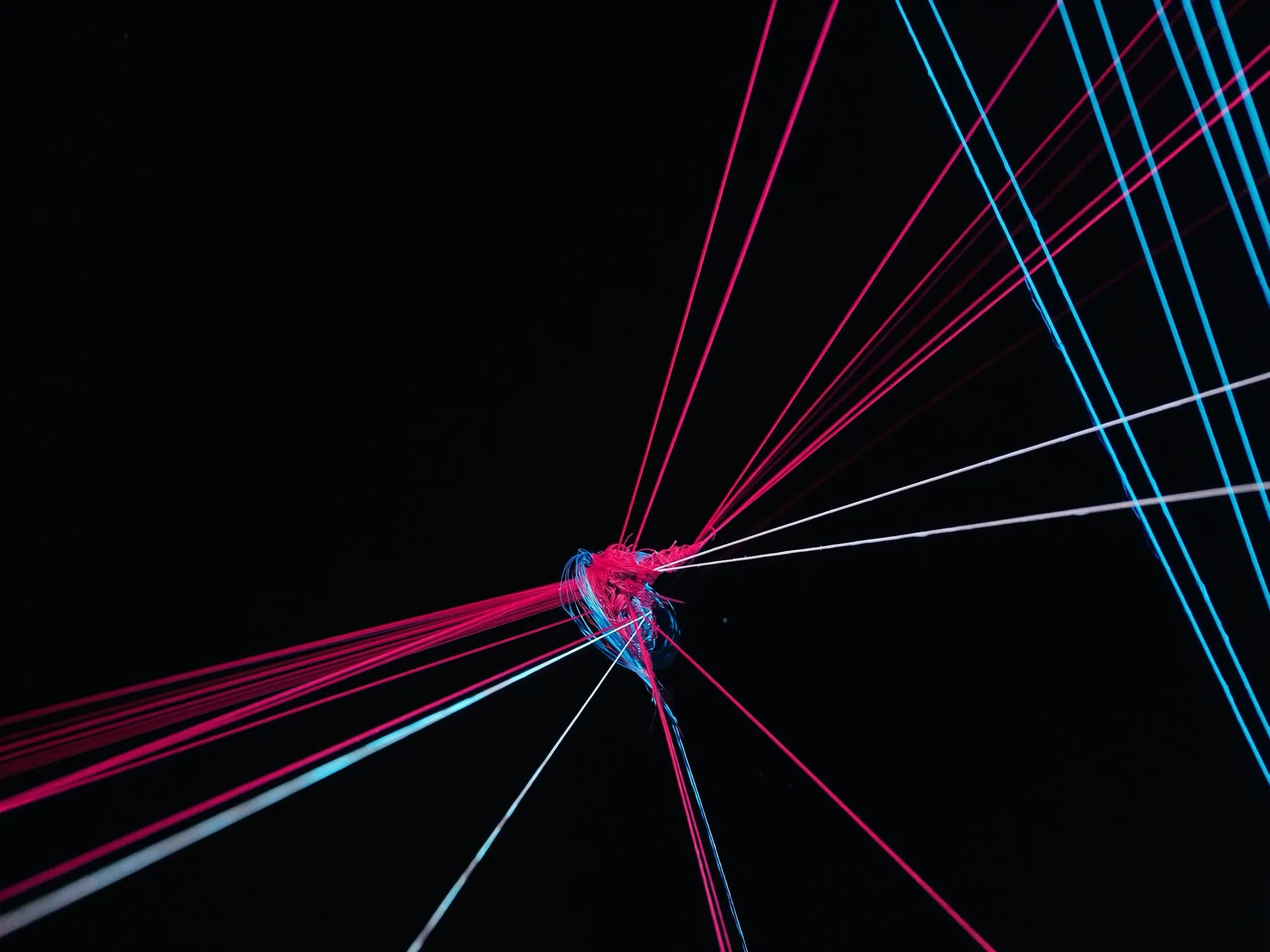 Dozens of thin threads pulled in different directions across a dark frame, tangled at the centre, lit hard from one side