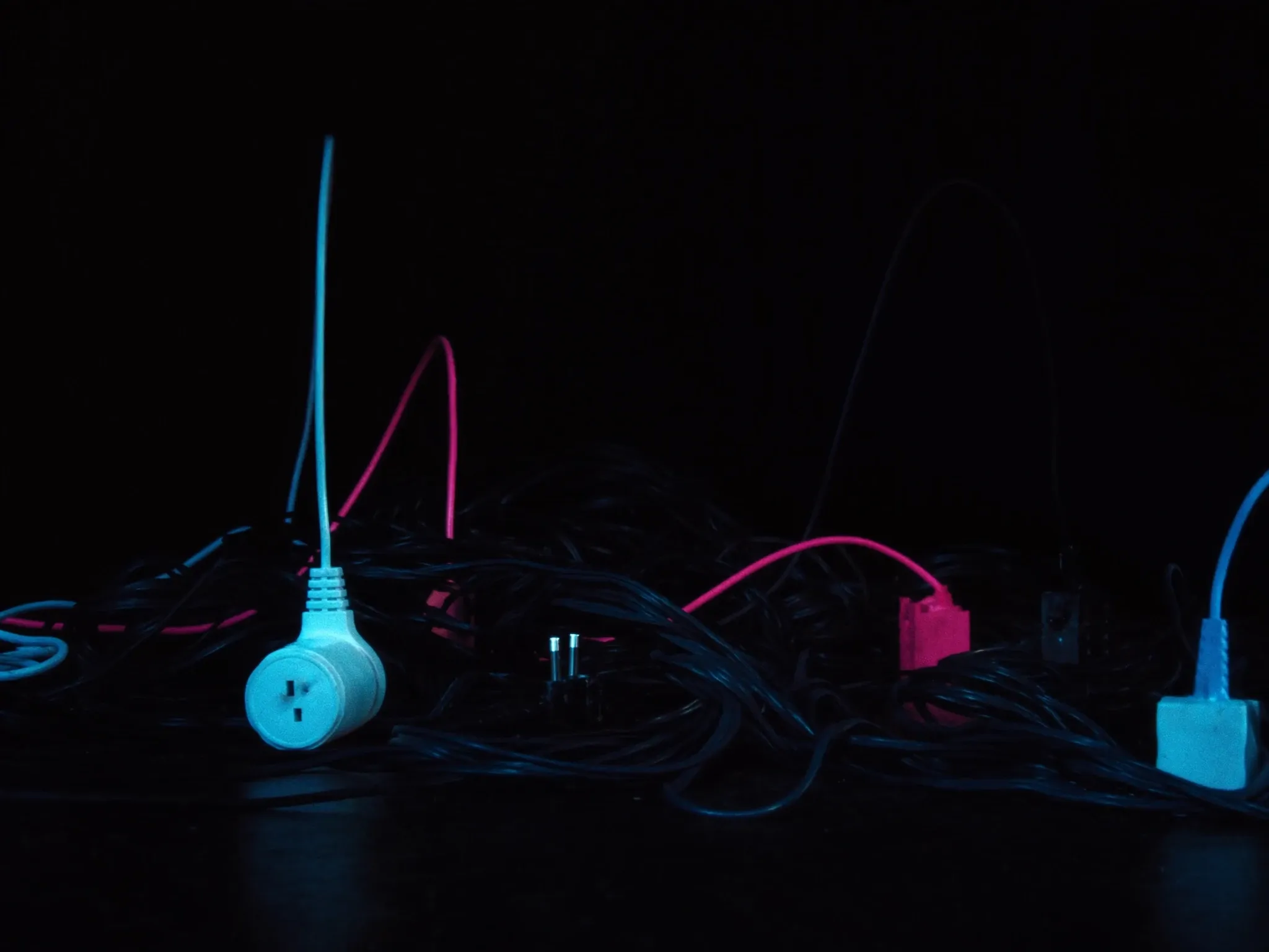 A tangle of mismatched cables and plugs on a dark floor, hard side light, no logos or lettering