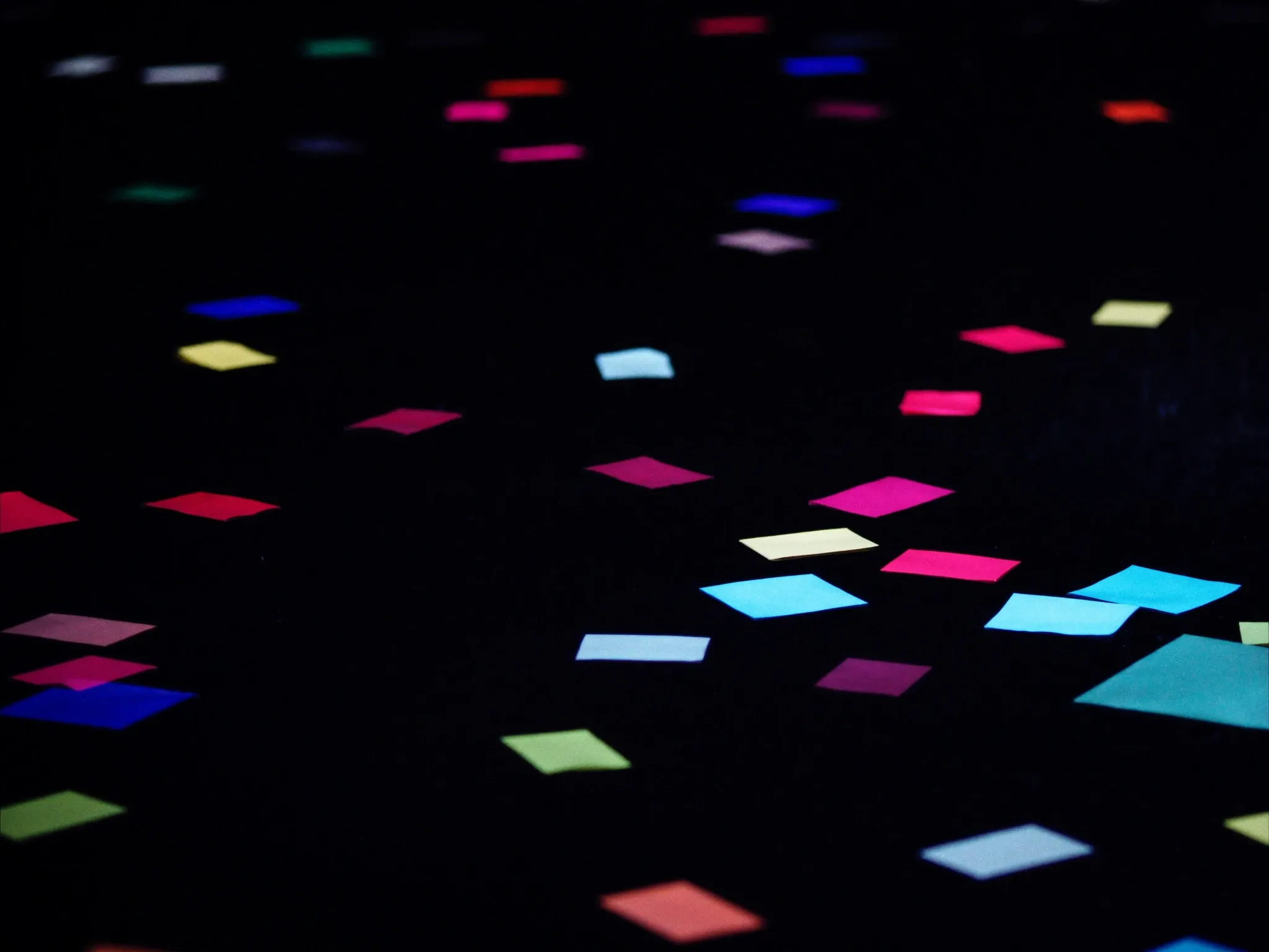 Scattered offcuts of coloured card in clashing colours on a dark surface, lit hard from one side