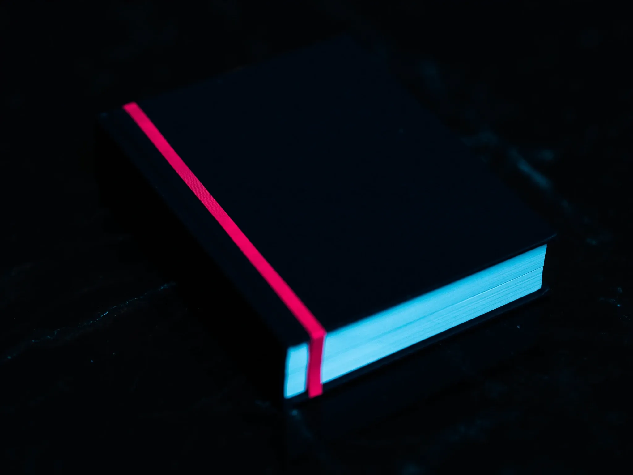 A heavy closed book bound in dark cloth with a thin pink ribbon marker, lying on black stone, soft light, blank cover