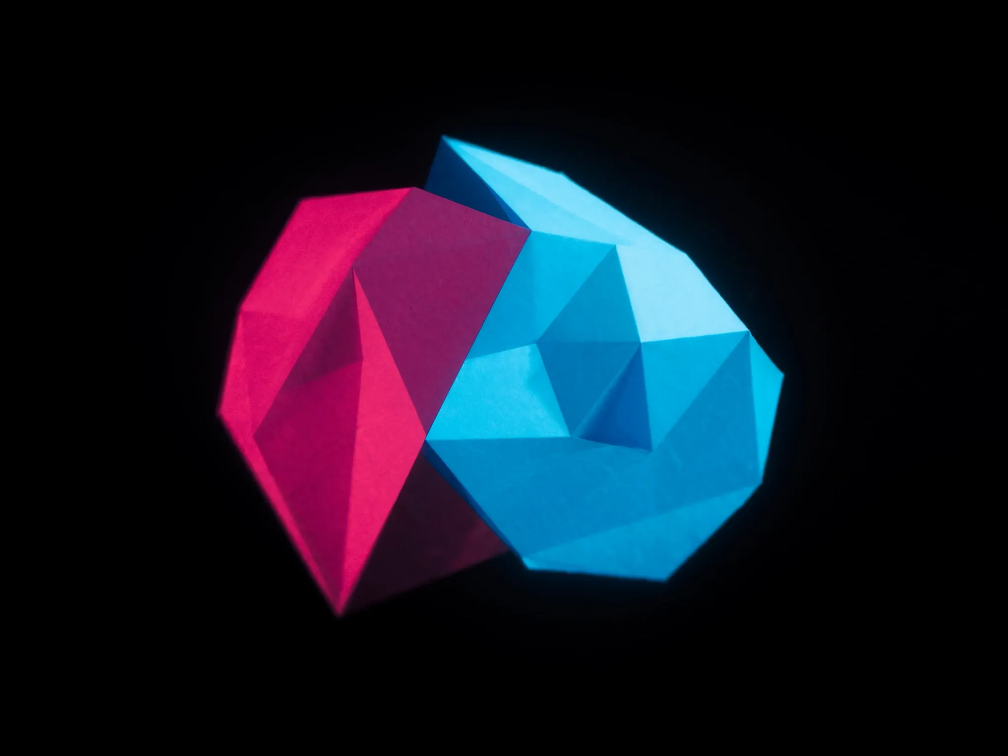 Interlocking geometric paper forms in pink and blue fitting together into one shape, studio light against black