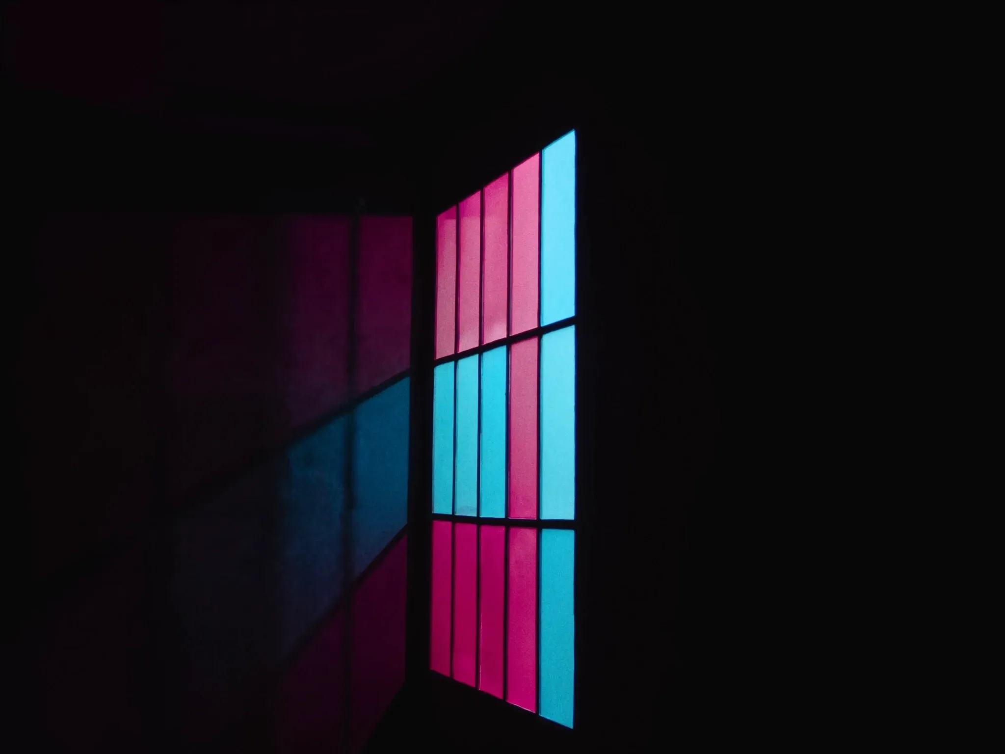 Light passing through a precise grid of glass panes, casting an ordered pattern of pink and cyan on a dark wall