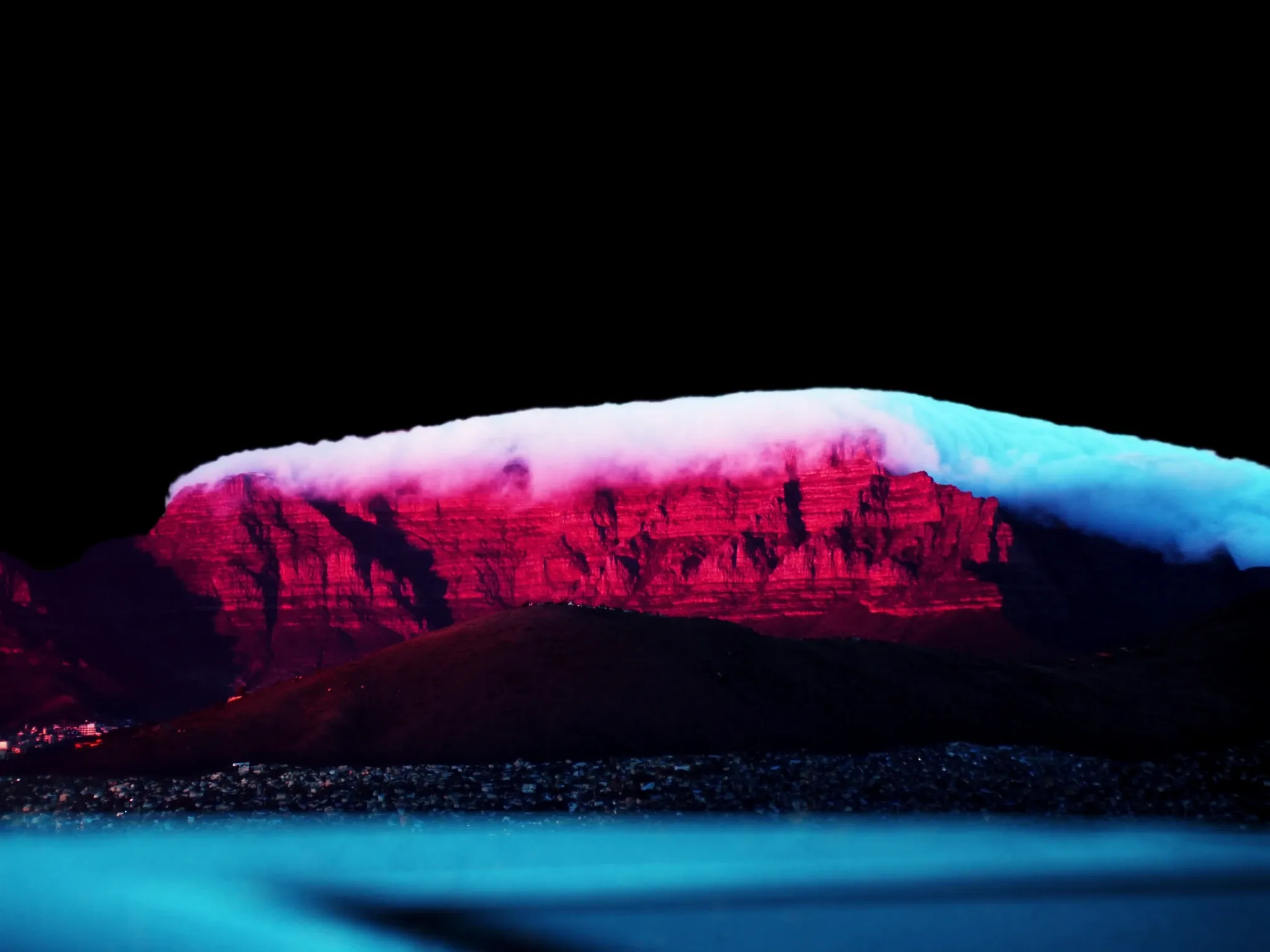 Table Mountain's long flat-topped plateau seen from across the bay at dusk, the white tablecloth cloud spilling over its edge and lit pink, the mountain clearly recognisable