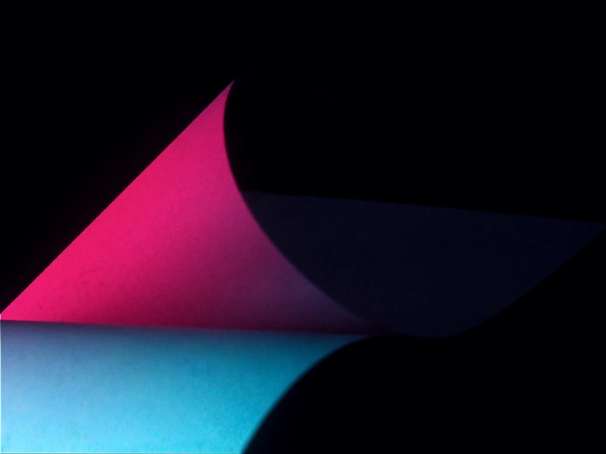 A single crisp fold in a sheet of heavy matte paper, one face lit pink and the other in deep shadow, against near-black