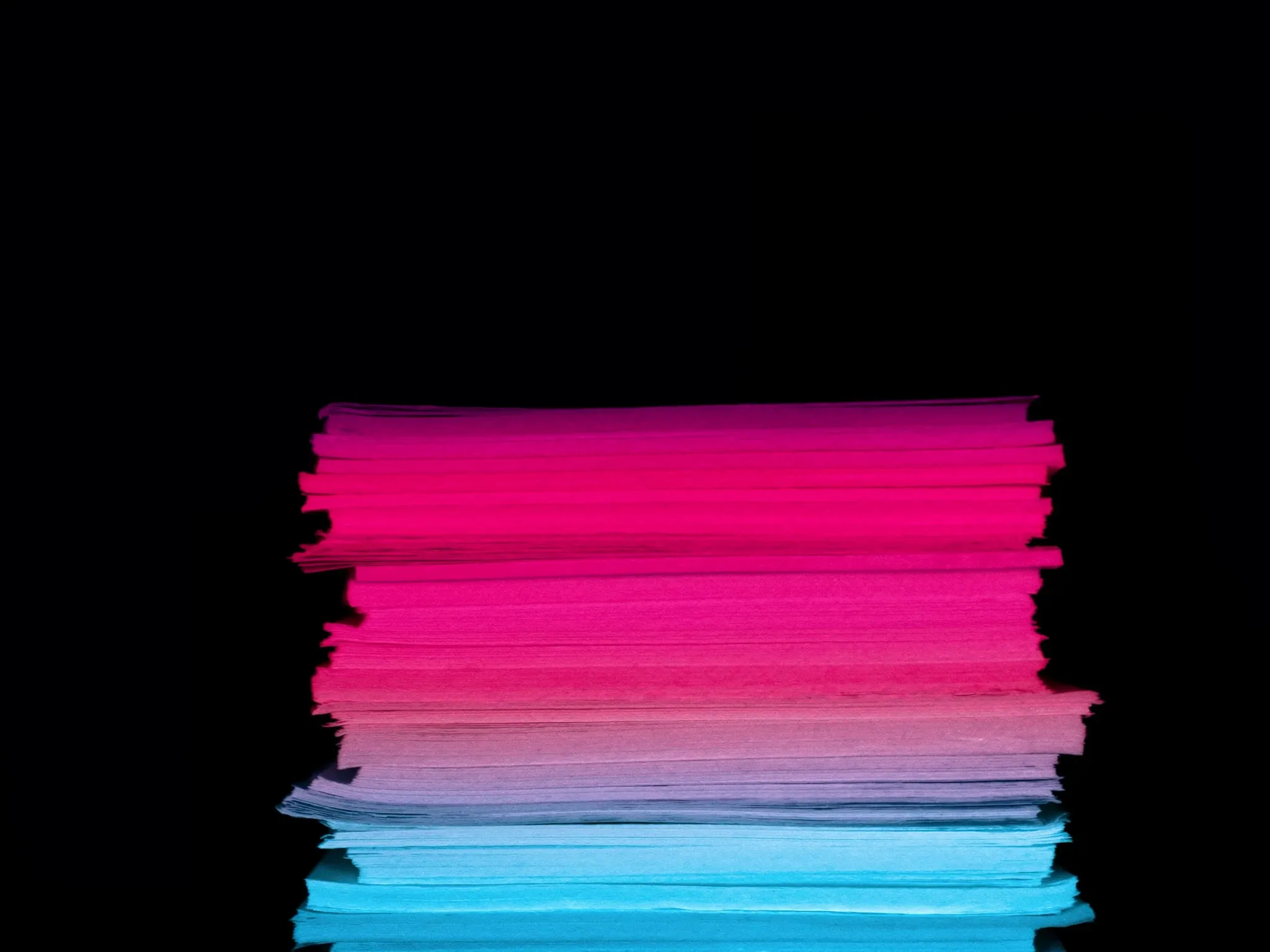 A stack of thick paper sheets in graded tones from deep magenta to pale pink, perfectly aligned, seen edge-on against black